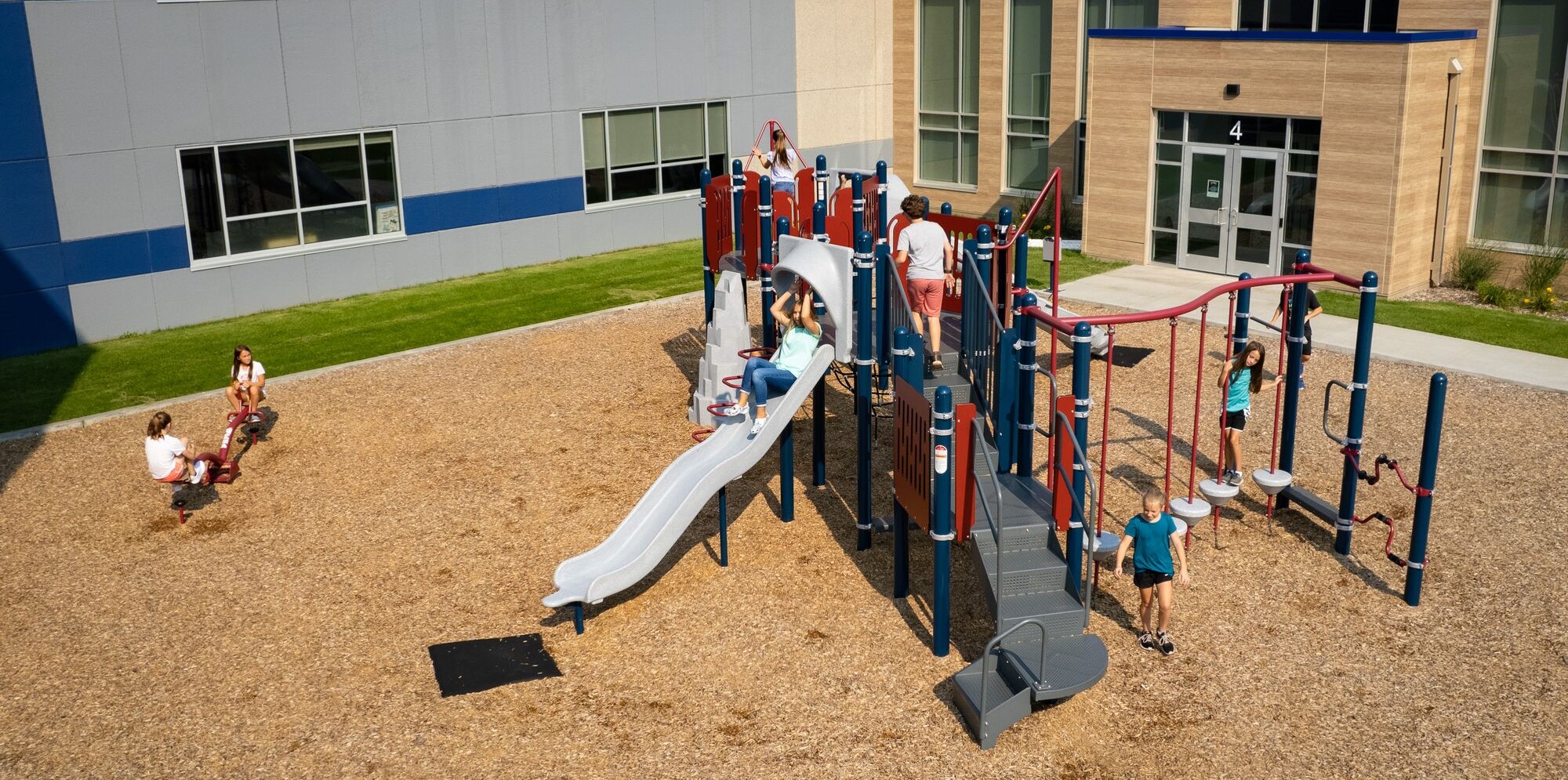 Wide shot of compact playground structure outside of a school-type building, with children playing on it