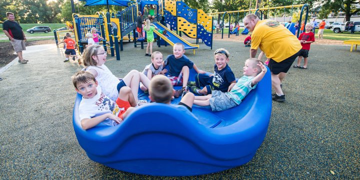 Smiling children sitting on an inclusive playground spinner while an adult pushes them. 