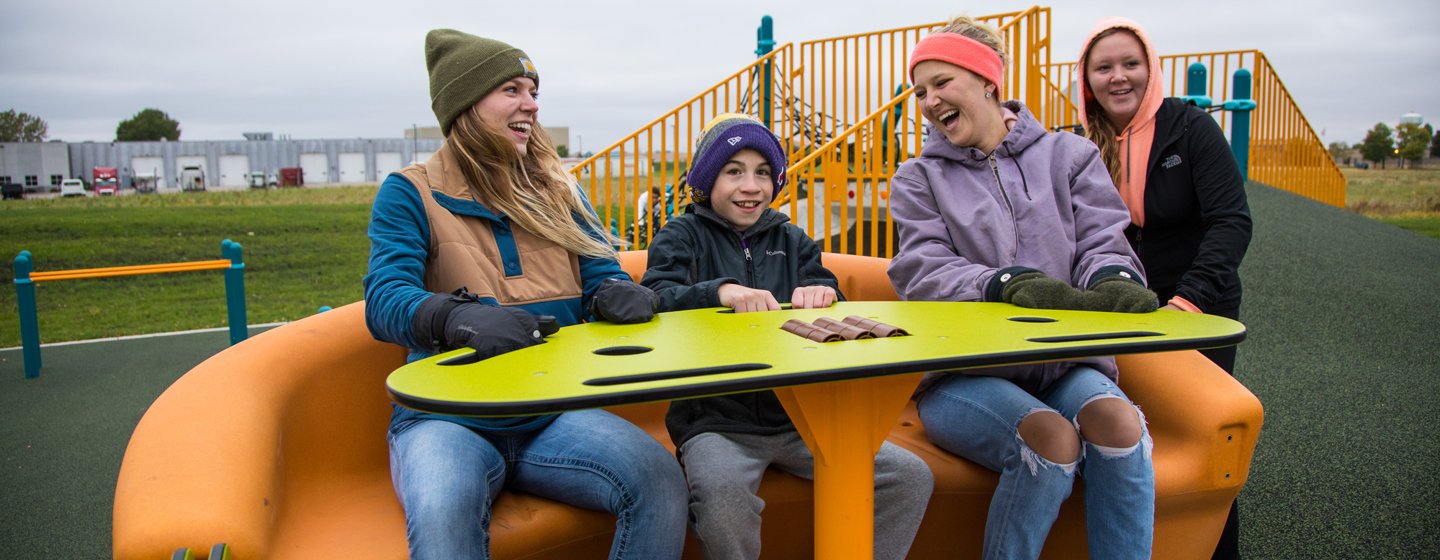Two adults and a child sit on the Sway Fun Glider at the Fallenstein Playground in North Mankato